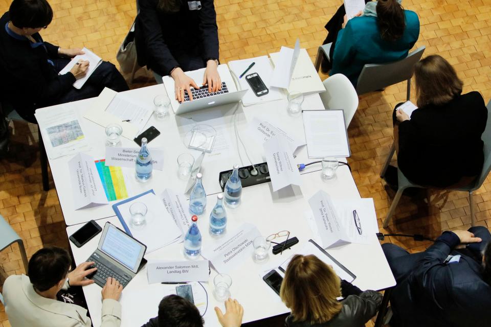 Viele Menschen an einem Gruppentisch mit Laptops und Schreibmaterialen. 
